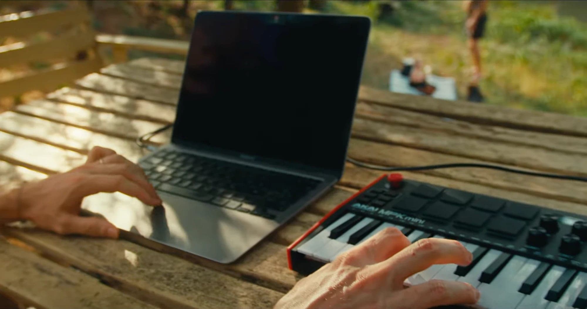 Hands on a laptop and a small MIDI keyboard on a wooden table outdoors