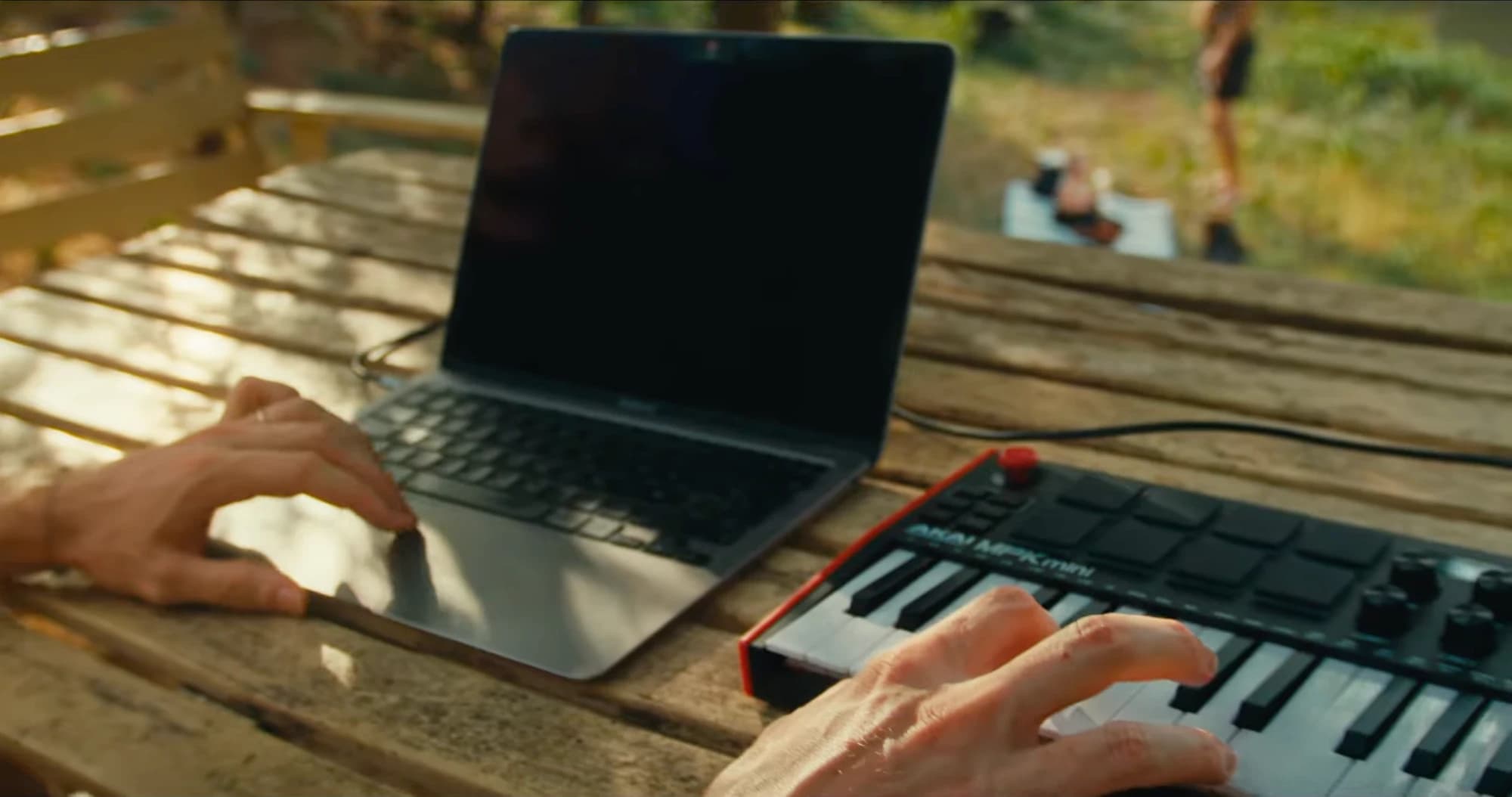 Hands on a laptop and a MIDI keyboard on a wooden table outdoors, mid creative challenge